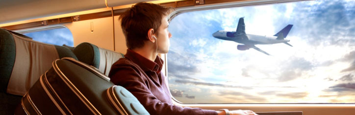 A young man traveling, seated with a suitcase in the seat beside him, watching out the window at an airplane taking off
